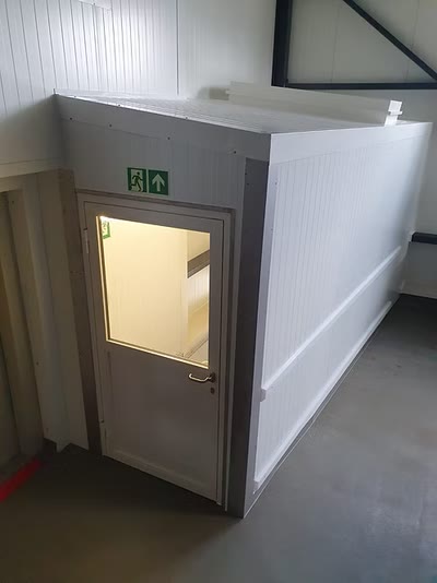 Eingebauter Raum mit Metalltür, Sichtfenster und Fluchtwegkennzeichnung in einer Gewerbehalle