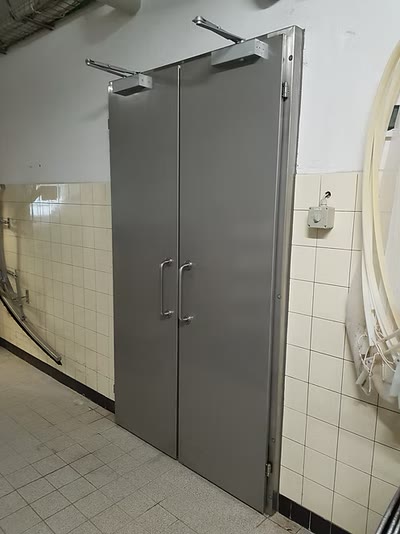 Zweiflügelige graue Metalltür mit obenliegenden Türschliessern und Edelstahlgriffen in einem geflieselten Betriebsraum
